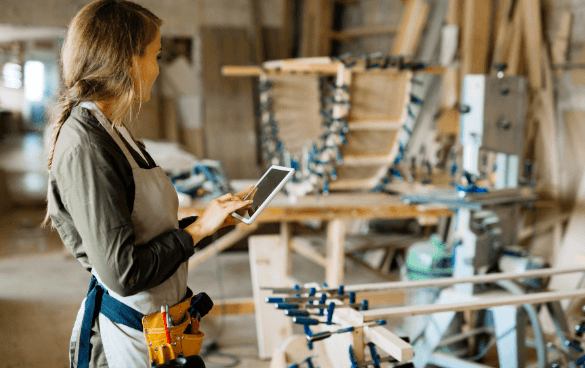 A woman in a woodworking shop wearing an apron and tool belt, holding a tablet, with woodworking tools and clamps in the background.