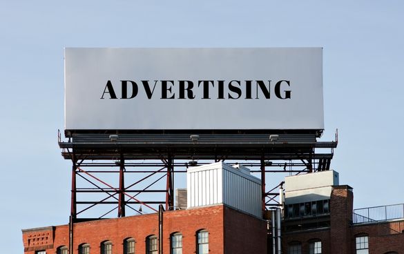 A billboard with the word 'ADVERTISING' above red brick buildings against a clear sky.