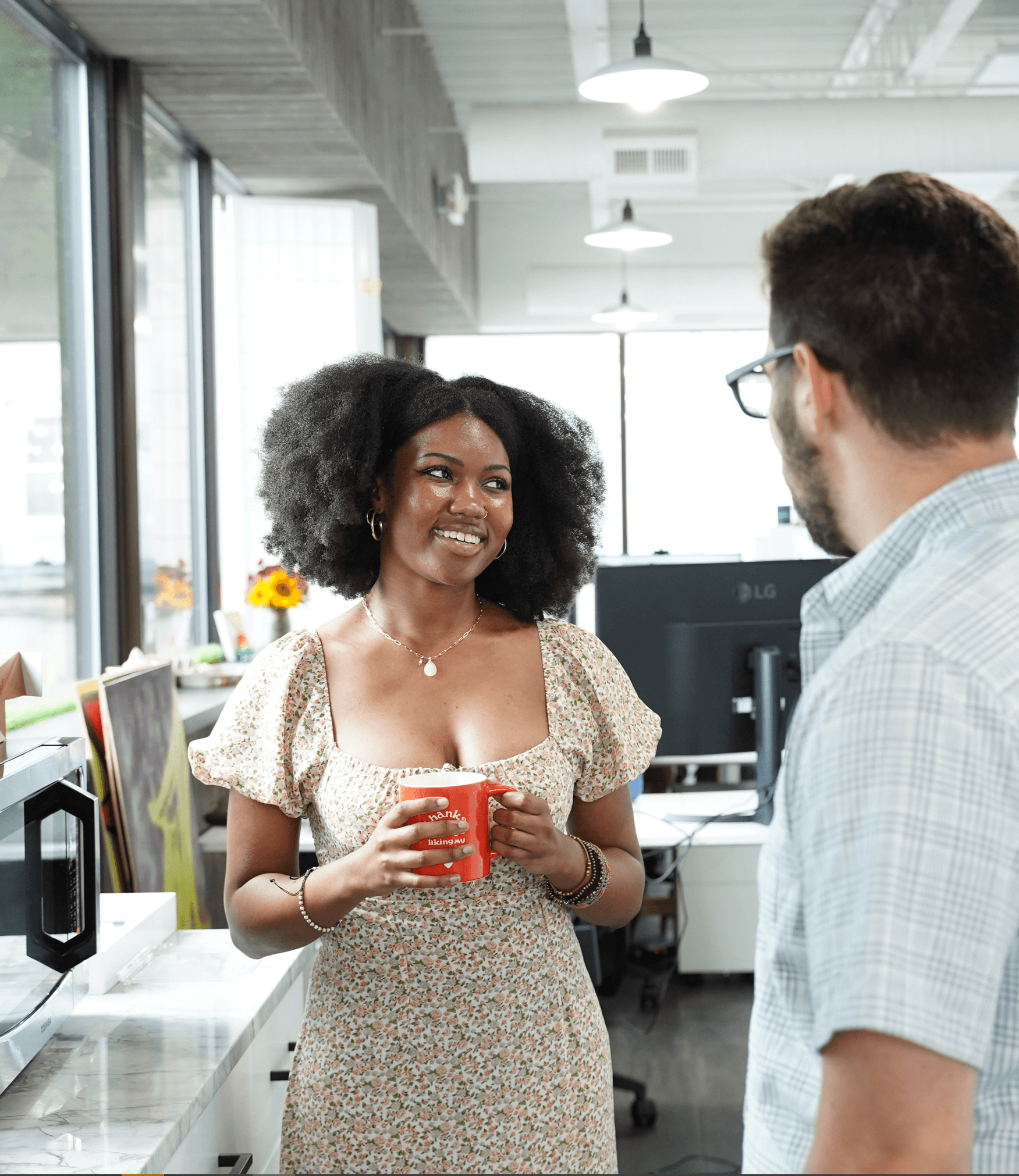 A smiling woman with an afro hairstyle holding a red mug in a modern office, engaging in a conversation with a man partially visible on the right.