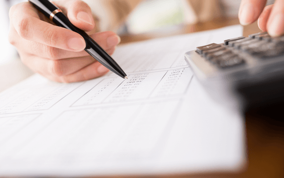 Close-up of a person's hands holding a pen and filling out a form with a calculator nearby, indicating financial paperwork or calculations.