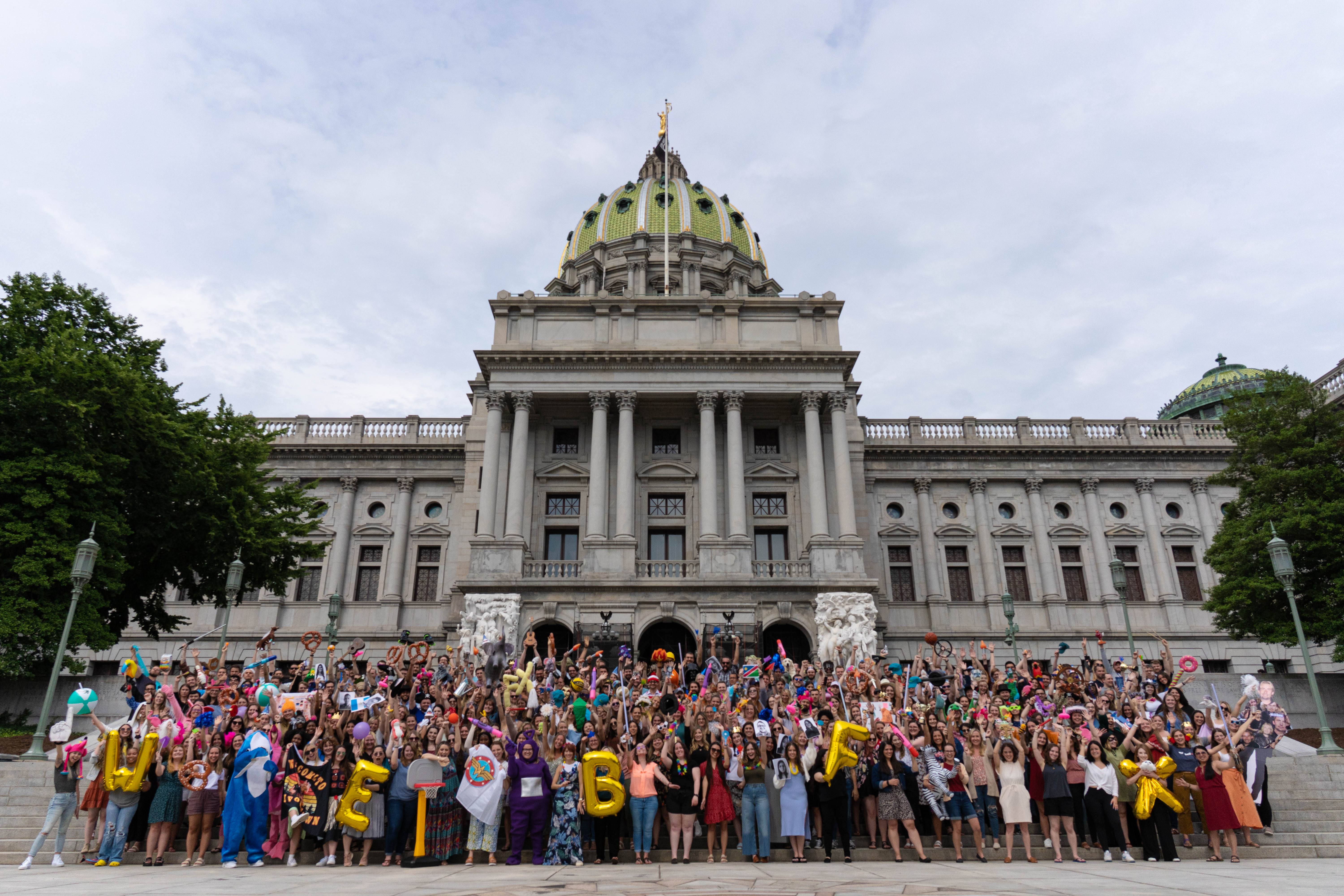 A diverse group of people gathered on the steps of a state capitol building, holding signs and colorful props, indicating a public event or demonstration.