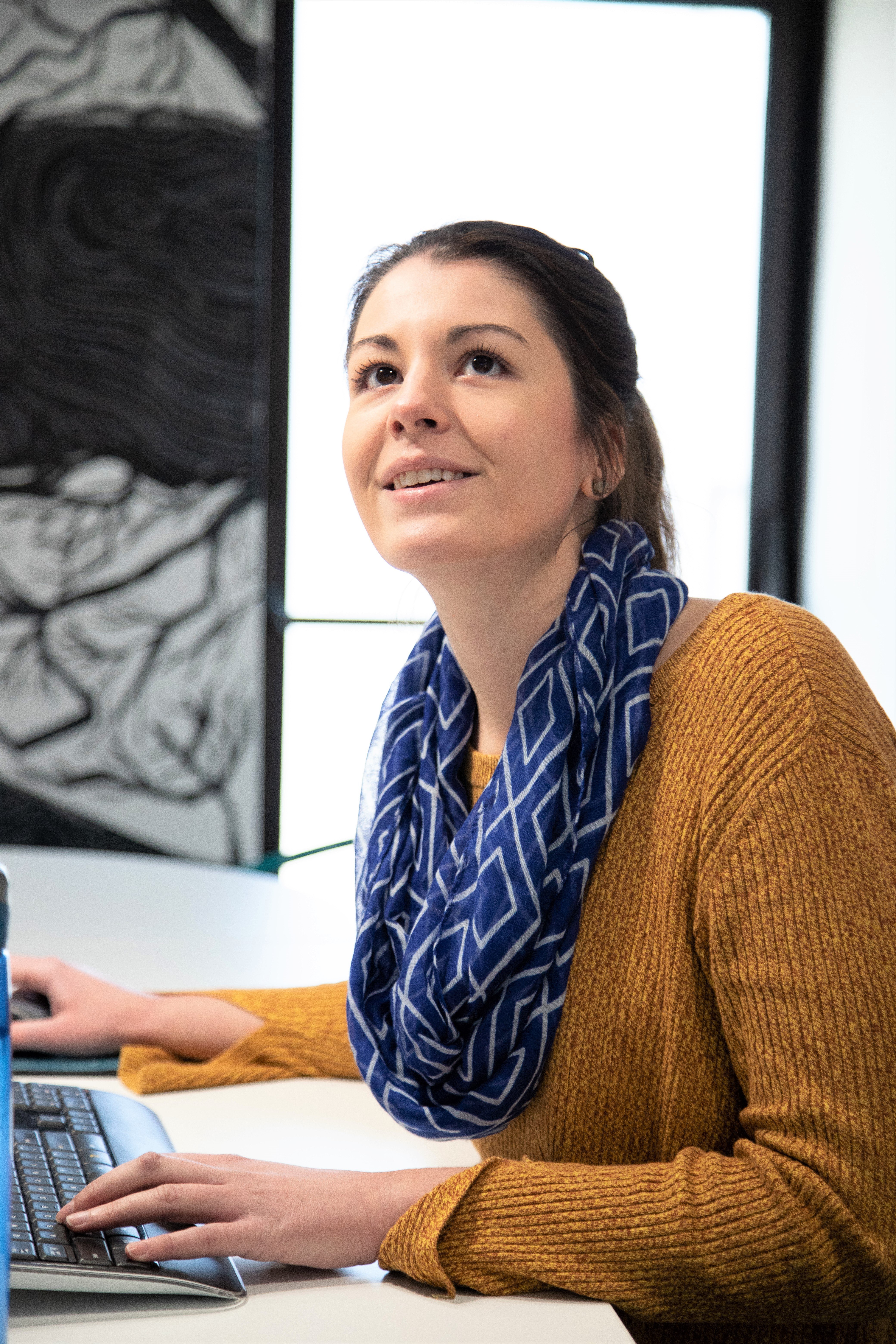 A woman in a mustard yellow sweater and blue scarf sits at a desk, looking up and smiling while using a laptop.