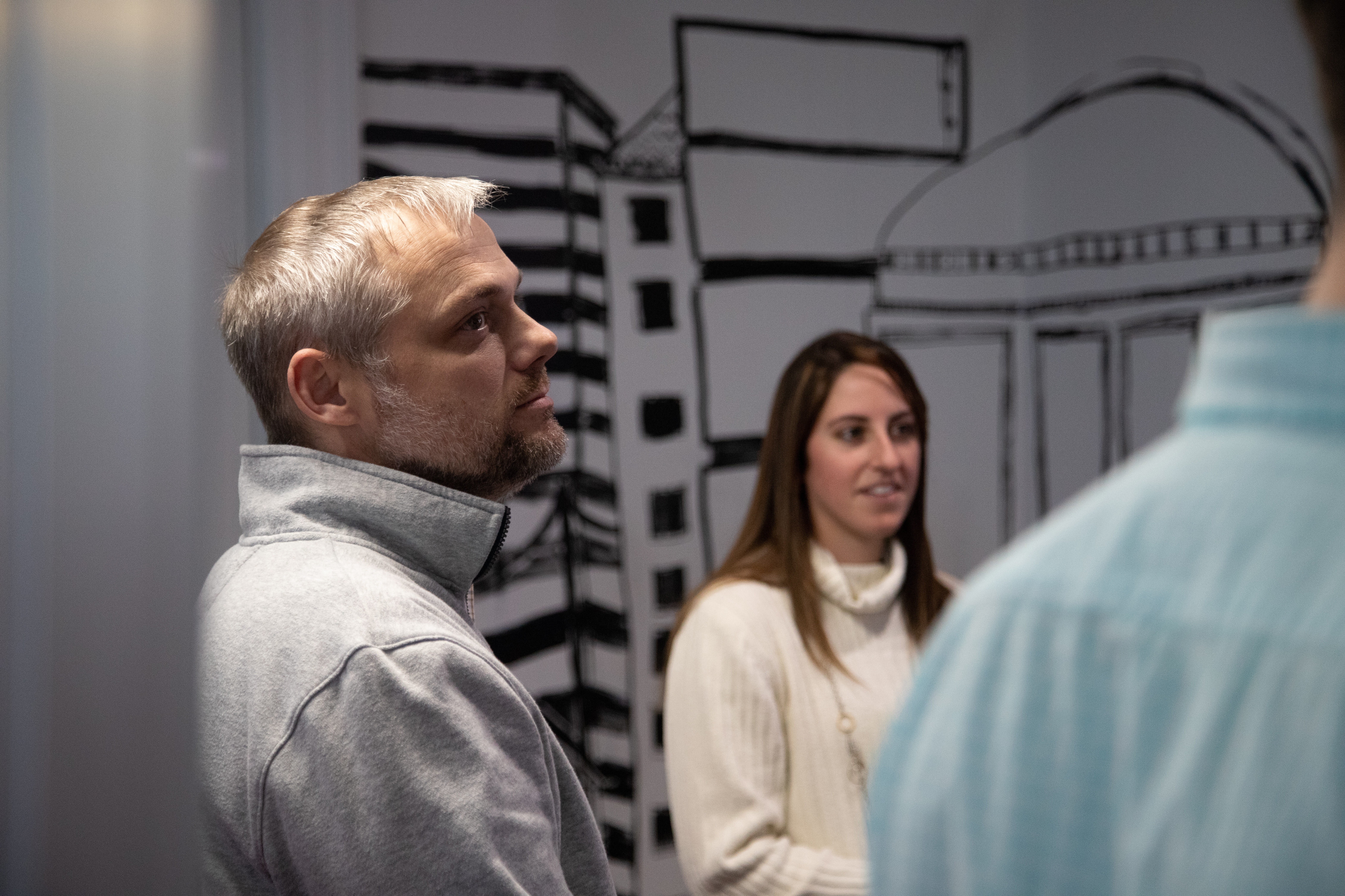 A man and a woman engaged in a conversation in a room with a wall drawing of buildings, with another person's back partially visible in the foreground.