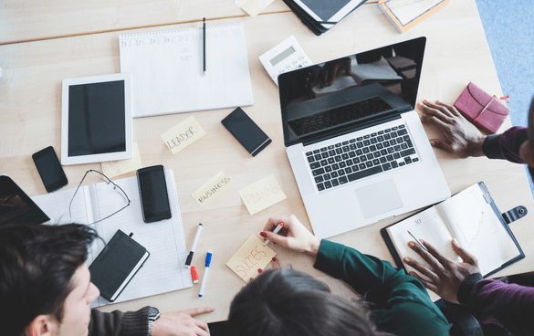 A top-down view of a collaborative workspace with three individuals, a laptop, digital tablet, smartphone, notebooks, pens, and sticky notes with words such as 'LEARN,' 'BUSINESS,' and 'SUCCESS.'