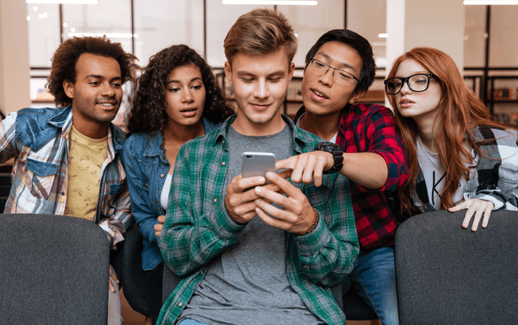 A diverse group of five young adults intently looking at a smartphone held by one of them in an indoor setting.