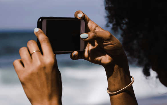 Person with curly hair taking a photo with a smartphone at the beach, with waves in the background.