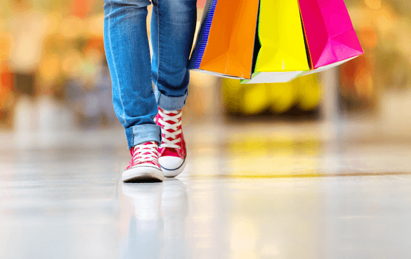 Person wearing blue jeans and red sneakers carrying multiple colorful shopping bags in a blurred shopping mall setting.