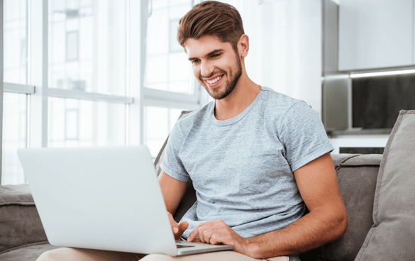 Smiling young man with a beard using a laptop while sitting on a couch in a bright, modern room.