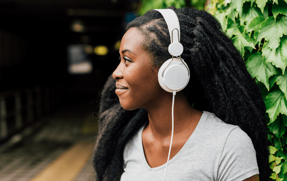 A content young woman with dreadlocks wearing a gray t-shirt and white headphones stands in front of a wall covered in green ivy.