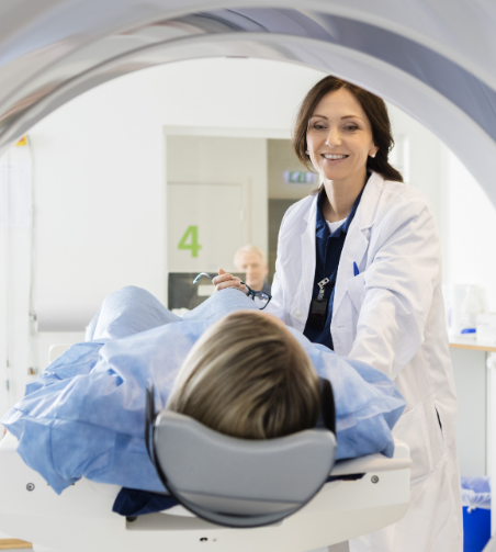 A smiling healthcare professional in a white lab coat stands beside a CT scanner, attending to a patient lying on the scanner bed, ready for a scan.