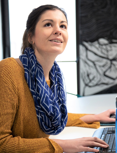 A woman in a mustard sweater and blue patterned scarf sitting at a desk, looking up and smiling slightly while using a computer keyboard.