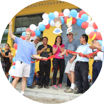 A man in a blue shirt is cutting a red ribbon with oversized scissors at a ribbon-cutting ceremony, surrounded by smiling people clapping, in front of a building decorated with balloons.