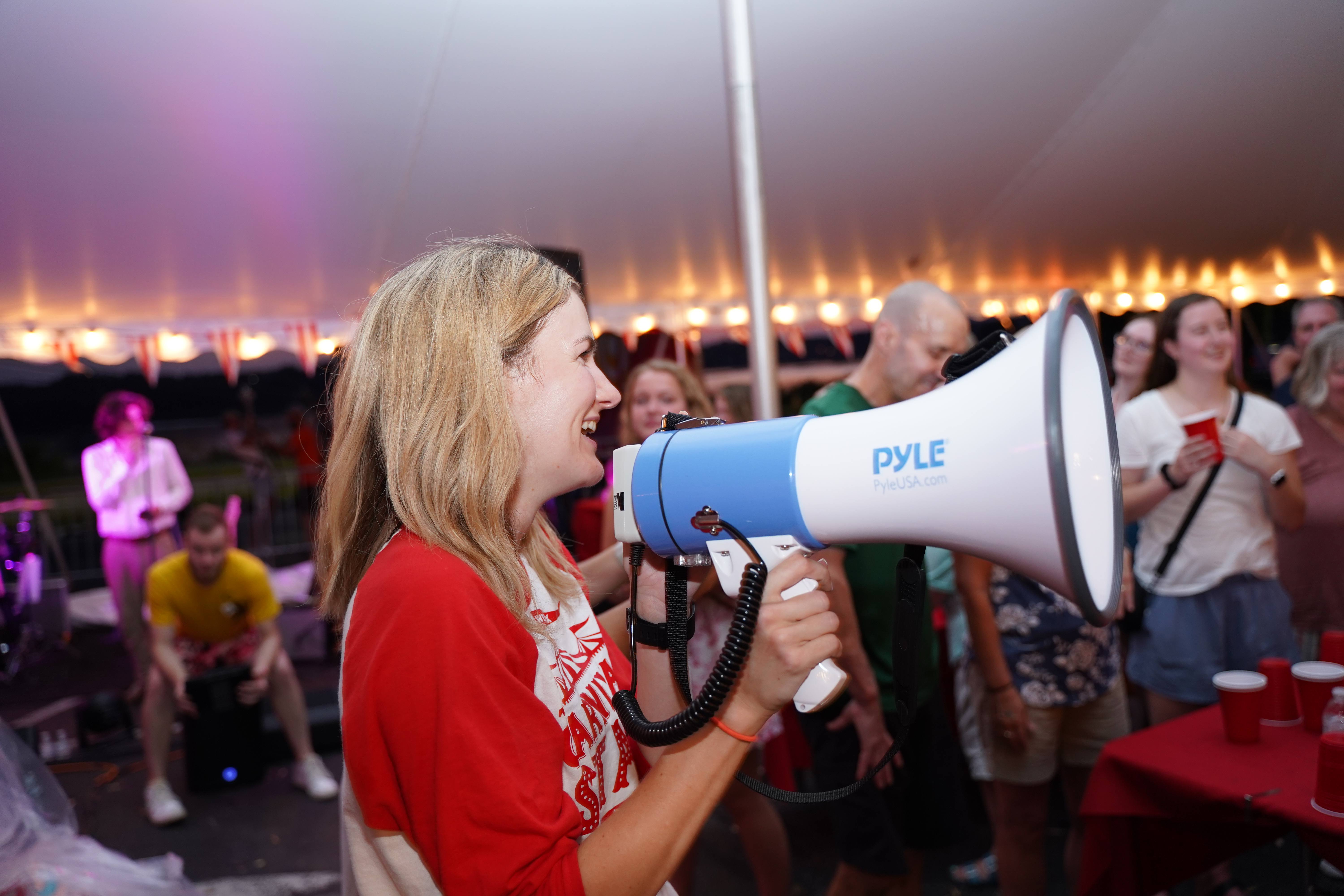 A woman joyfully speaking into a megaphone at a casual outdoor event with onlookers and a man playing a cajón in the background.