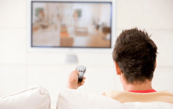 Person from behind sitting on a couch watching a mounted television screen and holding a remote control.