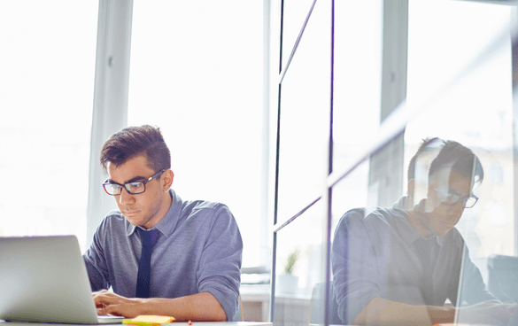 A focused young man with glasses working on a laptop at a desk in a brightly lit office, with a reflective surface creating a duplicate image beside him.