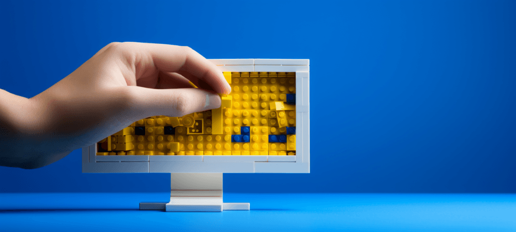 A hand placing a yellow LEGO brick onto a LEGO-built computer monitor with a white frame and stand against a blue background.