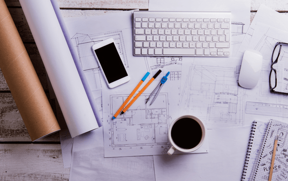 A professional workspace with architectural blueprints, a keyboard, mouse, smartphone, pencils, a ruler, a notebook, and a cup of coffee on a white desk.