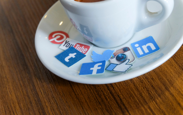 A white cup of coffee on a saucer with paper cutouts of social media logos including Pinterest, YouTube, Tumblr, Twitter, Facebook, and LinkedIn scattered on the saucer.