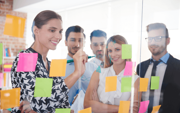 A team of five professionals collaborating around a glass wall covered with colorful sticky notes in a bright office setting.