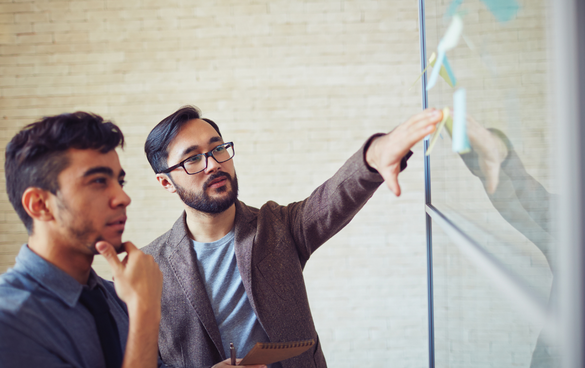 Two men collaborating in front of a glass board with sticky notes in an office setting.