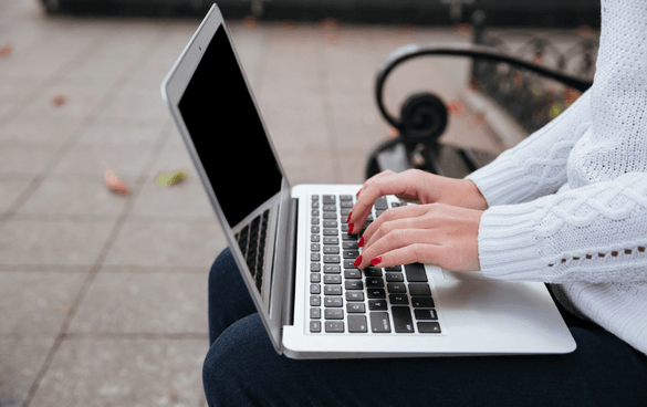 Person with red nail polish typing on a laptop while sitting on a park bench, with fallen leaves on the ground.