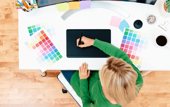 Top-down view of a graphic designer's workspace with a person using a drawing tablet, surrounded by color swatches, a coffee cup, and design tools.