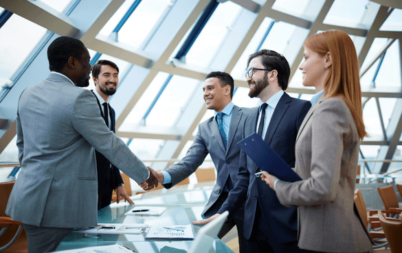 Five professionals in business attire engaging in a handshake and conversation in a modern office with a geometric glass ceiling.