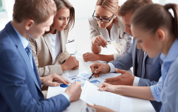 A group of five professionals in business attire engaged in a meeting, discussing over documents and charts on a table.