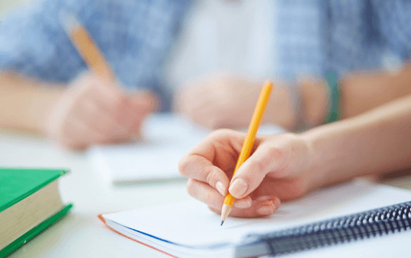 Two people writing in notebooks with pencils on a desk with books and notebooks, indicating a study environment.