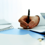 Close-up of a person's hand writing in a notebook with a pen, with a laptop and papers on the desk.