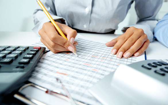 Person analyzing a printed spreadsheet with a pencil, with calculators and eyeglasses on the desk, indicating financial or accounting work.