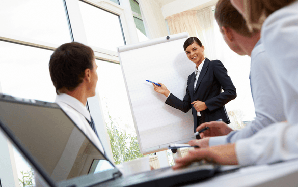 A woman in a business suit presenting at a flip chart to seated colleagues in a bright meeting room.