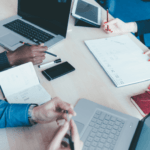 A group of professionals in a meeting with laptops, tablets, and documents on a table, actively engaged in discussion and work.