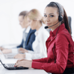 A woman in a red shirt with a headset working on a laptop at a call center, with colleagues in the background also wearing headsets and working on computers.