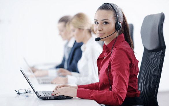 A woman in a red shirt with a headset working on a laptop at a call center, with colleagues in the background also wearing headsets and working on computers.