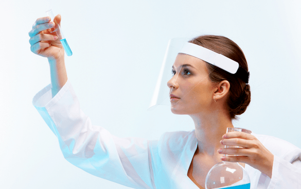 A female scientist in a lab coat and face shield examining a test tube with blue liquid while holding a flask with clear liquid.