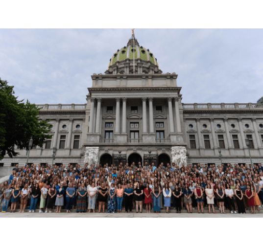 A large group of people posing for a photo on the steps of a grand capitol building with a green-domed roof.