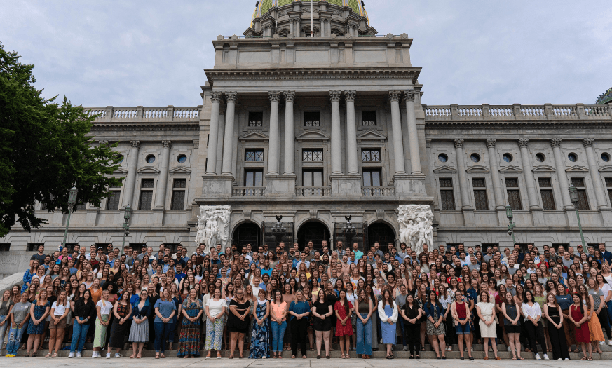 A large group of young adults posing for a group photo on the steps of an ornate government building with a domed roof and columns.