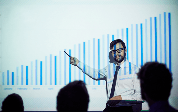 A man in glasses and business attire presenting a monthly data bar chart to an audience.