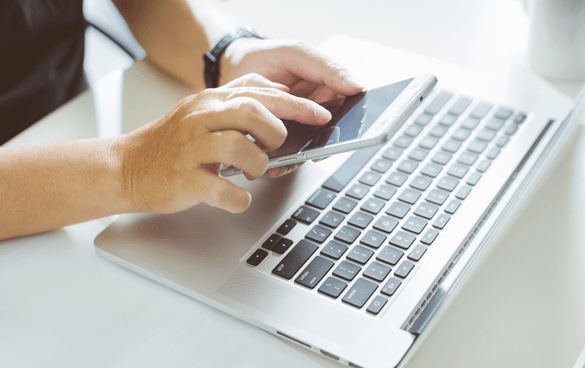 A person's hands with a watch on the left wrist, using a smartphone and interacting with a laptop's trackpad on a white surface.