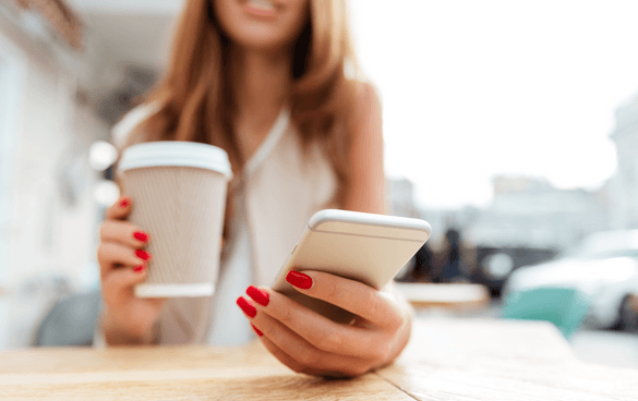 A woman's hands holding a disposable coffee cup and a smartphone, with red nail polish and a blurred outdoor background.