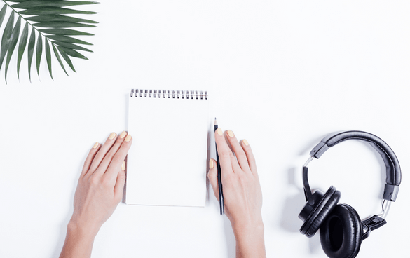 A pair of hands on a white surface, one holding a pen over a blank spiral notebook, with a pair of black headphones to the right and a green palm leaf in the top left corner.