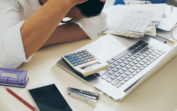 Person resting their head on their arm at a desk with a laptop, calculator, smartphone, and various papers indicating a state of stress or exhaustion.