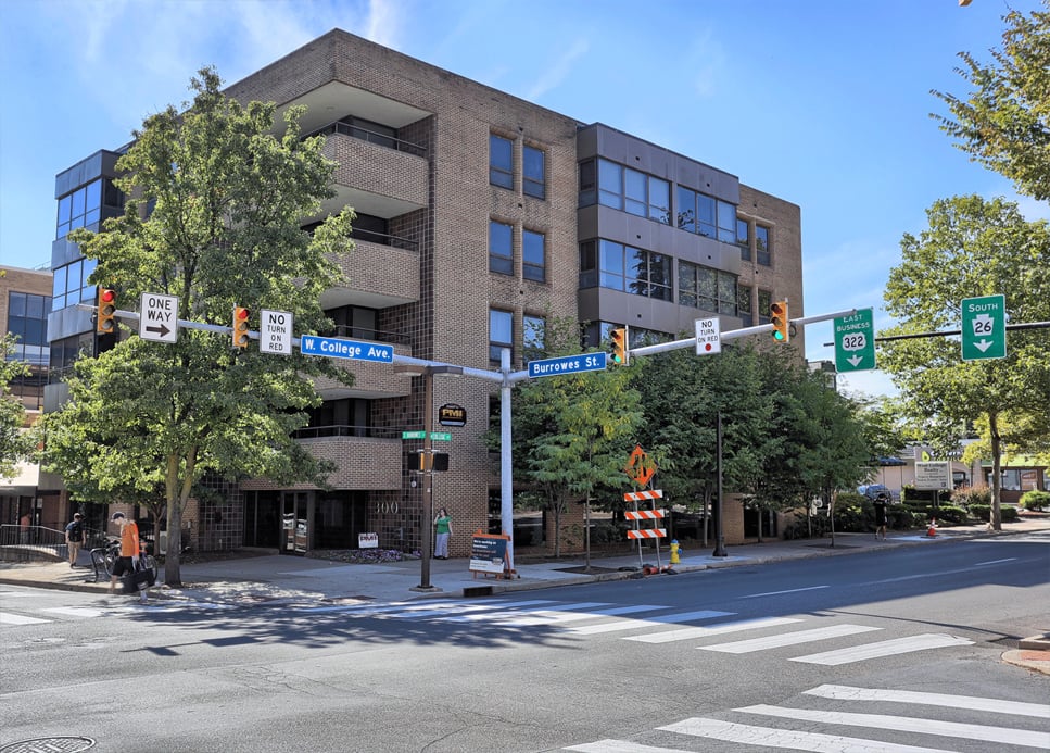 Street intersection with traffic lights and road signs at W. College Ave and Burrowes St, with directional signs to EAST 322 and SOUTH 26, pedestrians on the sidewalk, and a multi-story brick building on a sunny day with a clear blue sky.