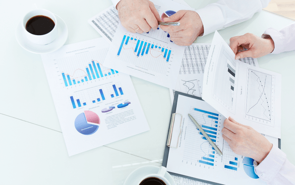 Two people reviewing and discussing printed data charts and graphs on a table with pens and coffee cups, indicating a business analysis or meeting.