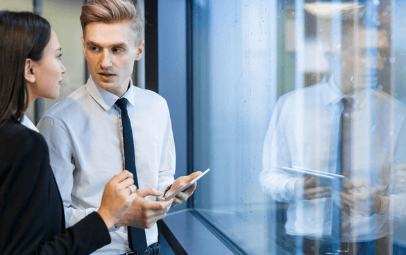 Two professionals in business attire, a man holding a smartphone and a woman, having a conversation near a glass window in an office setting.