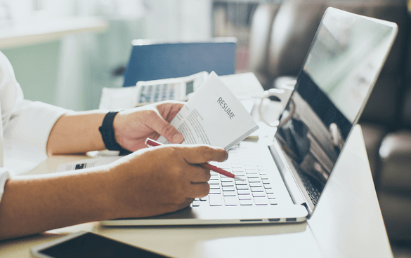 Person holding a resume while working on a laptop at a desk.