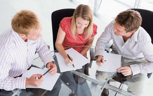 Three professionals, two men and one woman, are seated around a glass table in an office, engaged in a meeting with documents and pens in hand.