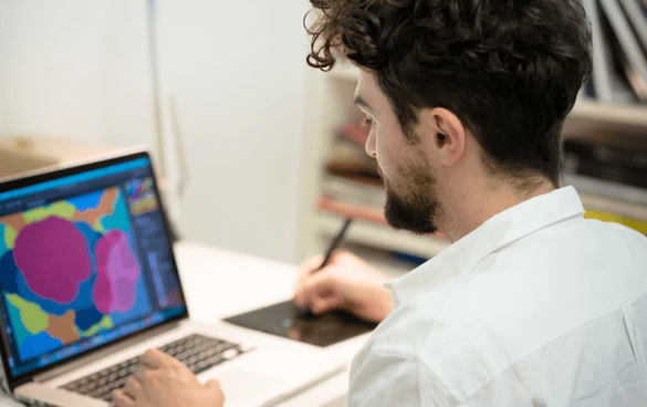 A man with curly hair working on a laptop with a colorful abstract design on the screen while using a digital pen on a graphics tablet.