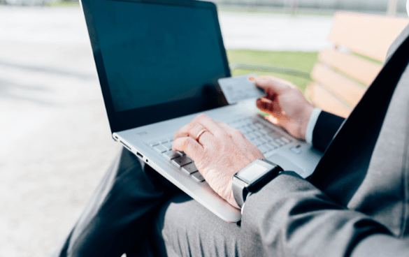 Person in a suit making an online purchase on a laptop while holding a credit card, with a smartwatch on their wrist, seated outdoors.